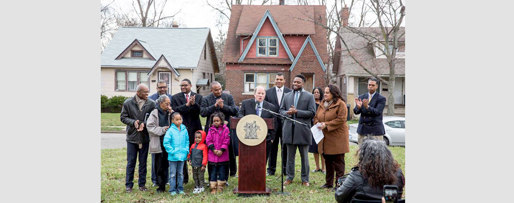Photograph of a man standing at a podium speaking into a microphone with several adults and children standing nearby; three single-family detached houses are in the background.