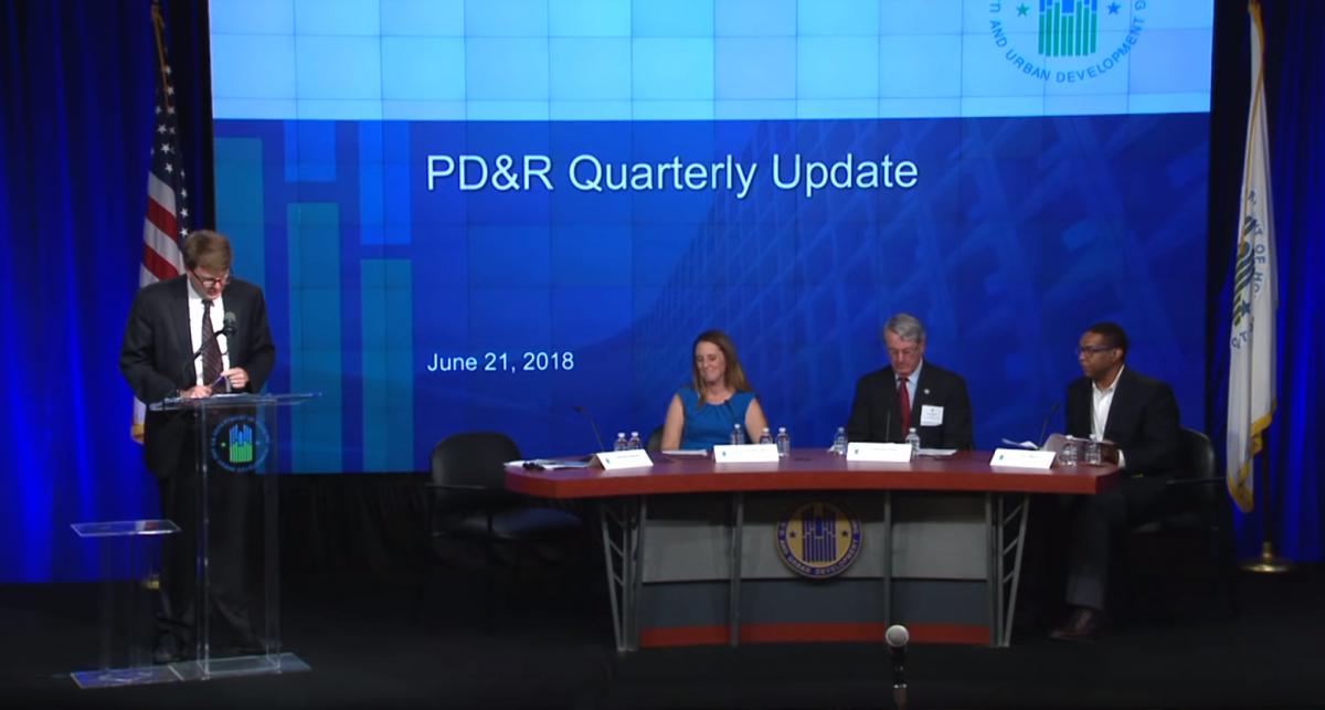 Todd Richardson, general deputy assistant secretary for PD&R, stands at a podium next to three panelists seated at a table. A screen with the words "PD&R Quarterly Update" is visible in the background. 